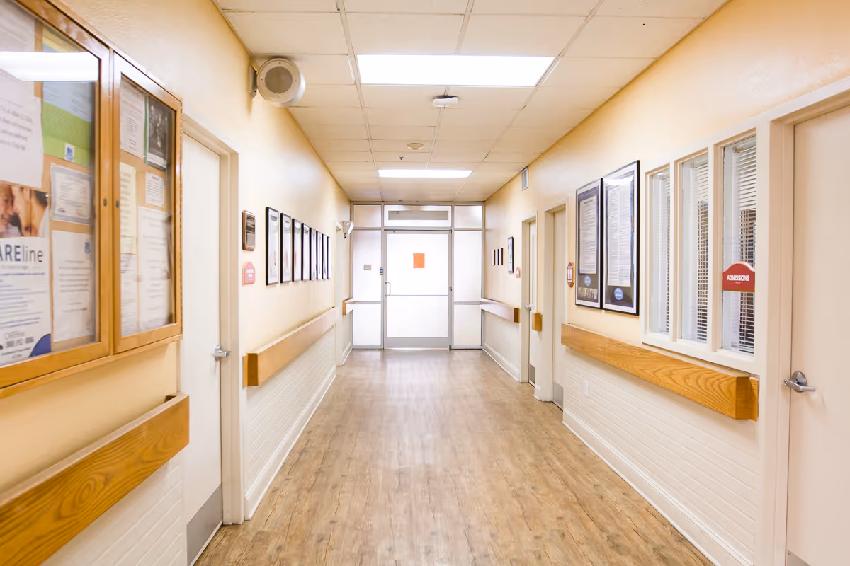 A clean, well-lit hallway in a care and rehab center with light yellow walls, wood handrails, framed notices and documents on the walls, and a wooden floor leading to a glass door at the end.