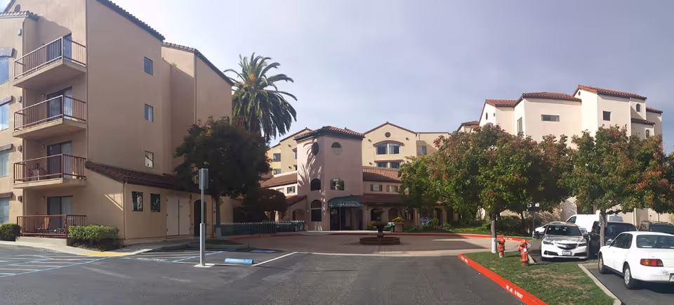 Front exterior of the Cadence Millbrae senior living complex with a driveway, fountain, parked cars, and multi-story stucco buildings with balconies and landscaping.