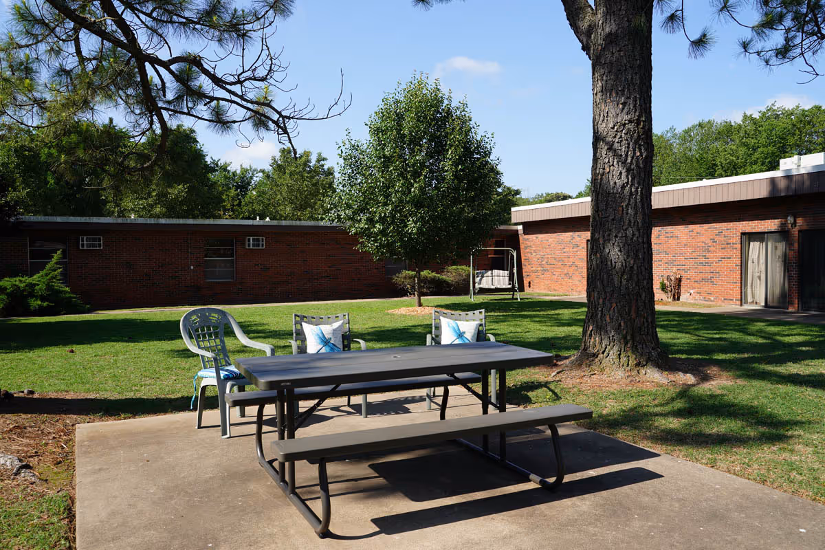 Outdoor patio area at Stilwell Nursing & Rehab featuring a picnic table with attached benches and three chairs with cushions, set on a concrete slab surrounded by grass, trees, and a brick building in the background under a clear blue sky.