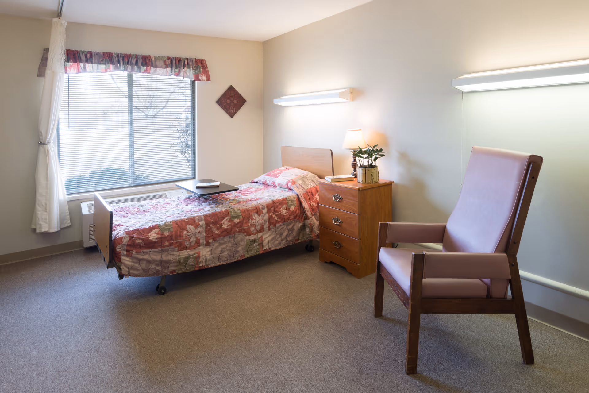 A tidy single-occupancy bedroom with a bed, nightstand and lamp, an armchair, and a window with blinds.