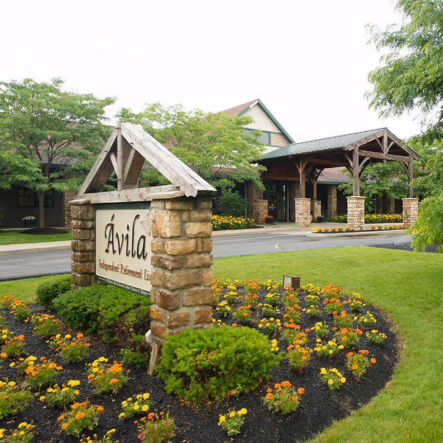 Entrance to Avila Retirement Community featuring a stone sign with the community name, surrounded by a landscaped garden with colorful flowers and green shrubs. In the background, there is a covered driveway supported by stone pillars and wooden beams, with trees and a building visible.