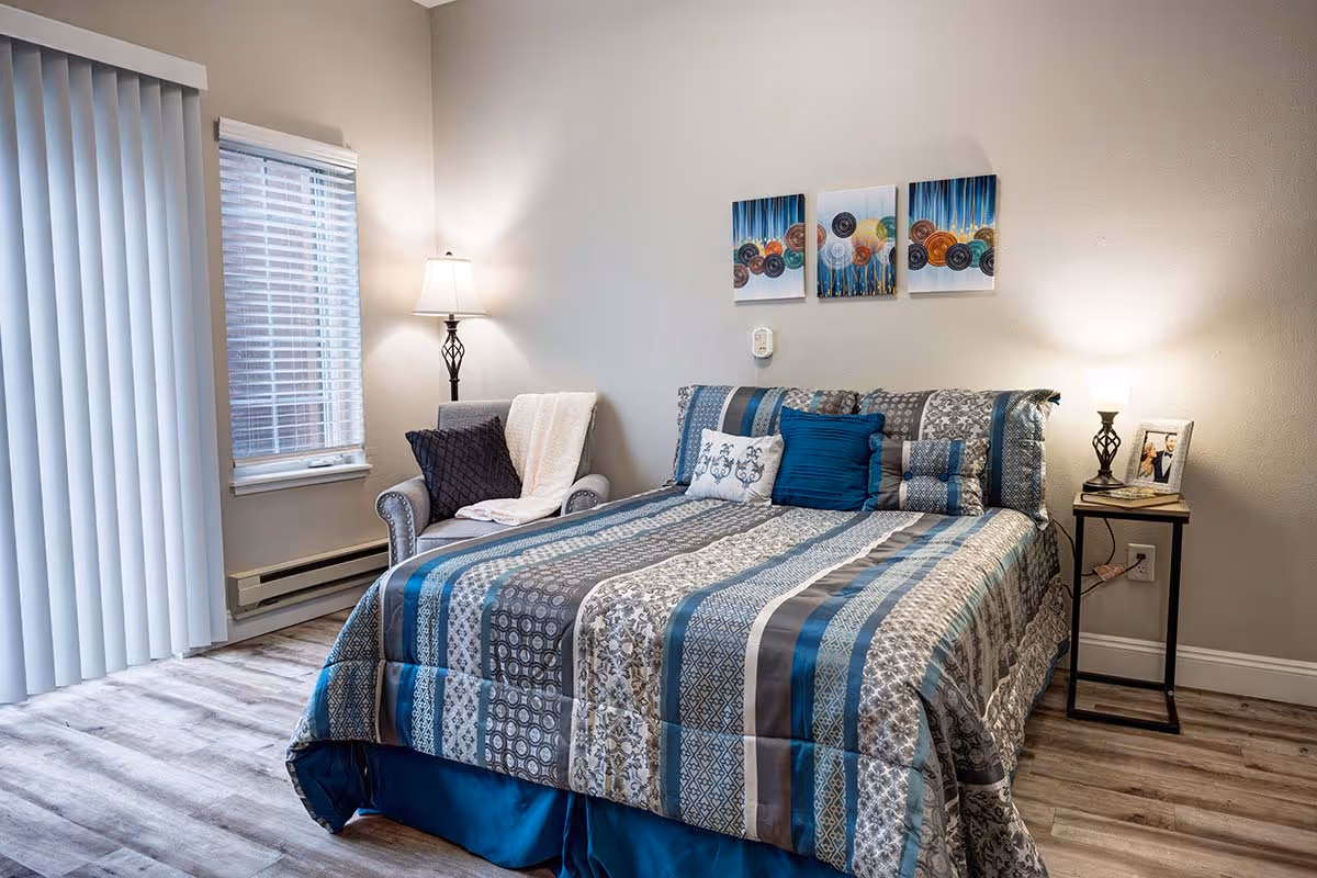 A cozy bedroom featuring a bed with a blue and gray patterned comforter and multiple pillows. To the left of the bed is a cushioned armchair with a throw blanket and pillow. There is a window with blinds and vertical blinds covering a sliding door. On the right side of the bed is a small nightstand with a lamp and a framed photo. Three decorative paintings hang on the wall above the bed.