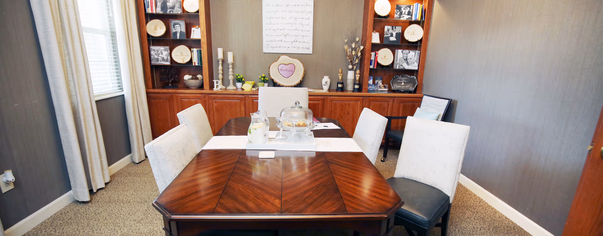 A dining room with a wooden table and six upholstered chairs. The table has a white runner and a tray with a glass cake dome and a pitcher. Behind the table is a built-in wooden cabinet with shelves displaying framed photos, candles, and decorative items. The room has gray walls, carpeted floor, and a window with light-colored curtains on the left side.