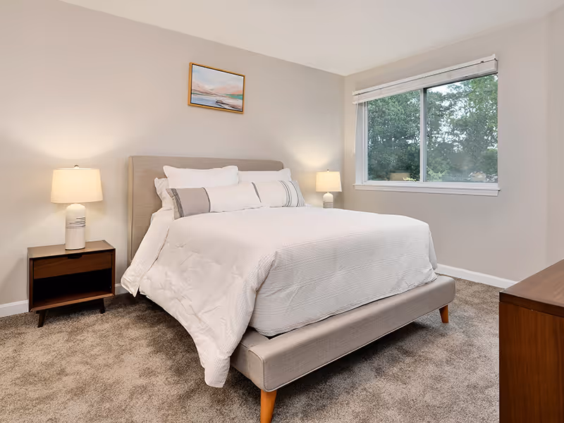 A neatly made bed with white bedding and multiple pillows in a bedroom. There are two bedside tables, each with a lamp. A window with a view of green trees is on the right wall, and a small framed painting hangs above the bed. The room has beige walls and carpeted flooring.