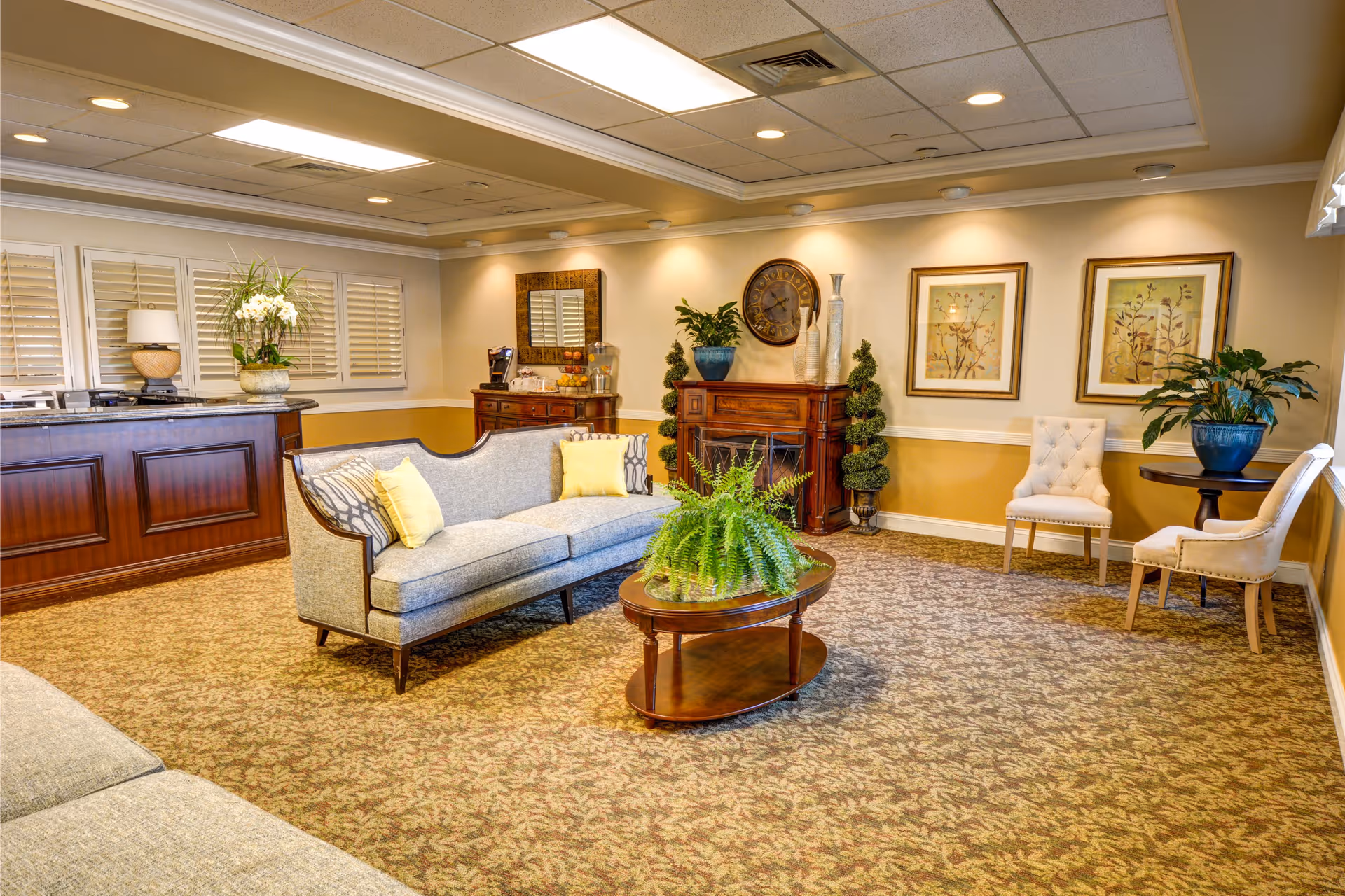 A cozy and well-lit living room area with a patterned carpet, a gray upholstered sofa with yellow and patterned pillows, a wooden coffee table with a green fern plant, two beige tufted chairs around a small round table with a potted plant, and a wooden sideboard with decorative items including a clock, vases, and framed artwork on the walls.