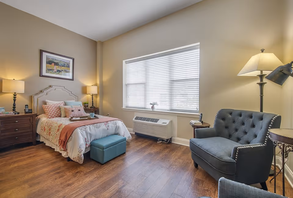 A cozy bedroom with a single bed adorned with multiple pillows and a floral bedspread. There are two bedside tables with lamps on either side of the bed. A blue upholstered armchair and a small round table with a lamp are positioned near a large window with blinds. The room has wooden flooring and neutral-colored walls.