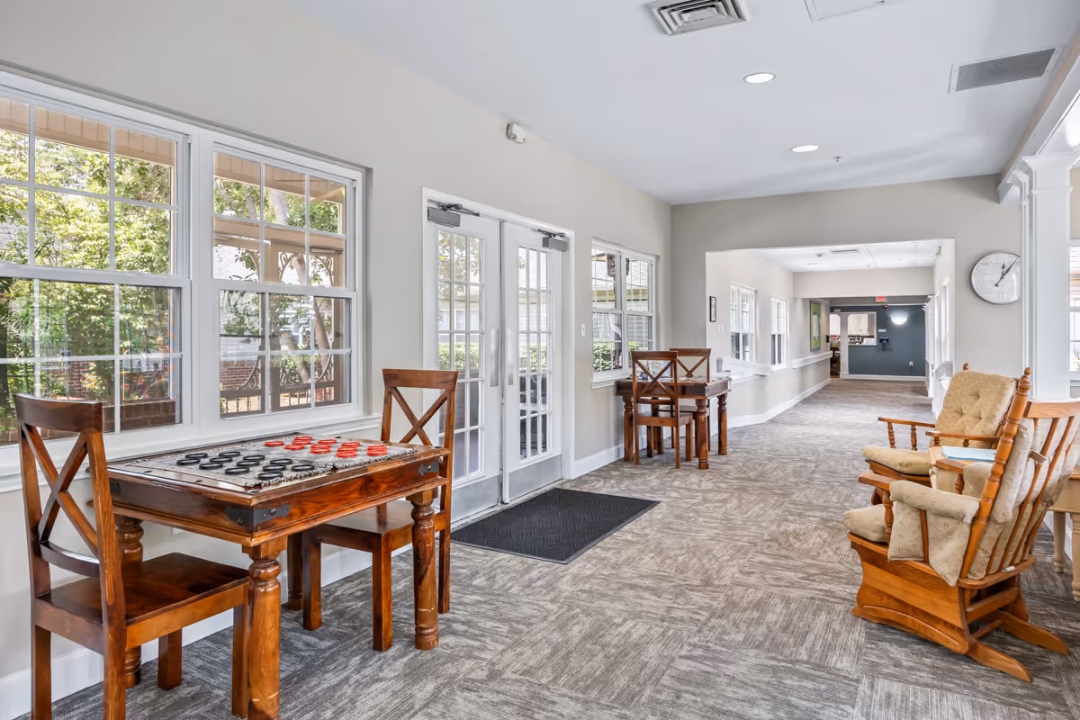 A bright hallway with large windows on the left side and double glass doors leading outside. There are two wooden tables with chairs, one table has a checkers game set up. On the right side, there are two cushioned wooden rocking chairs. The floor is carpeted and the walls are painted light gray. A clock is mounted on the wall near the rocking chairs.