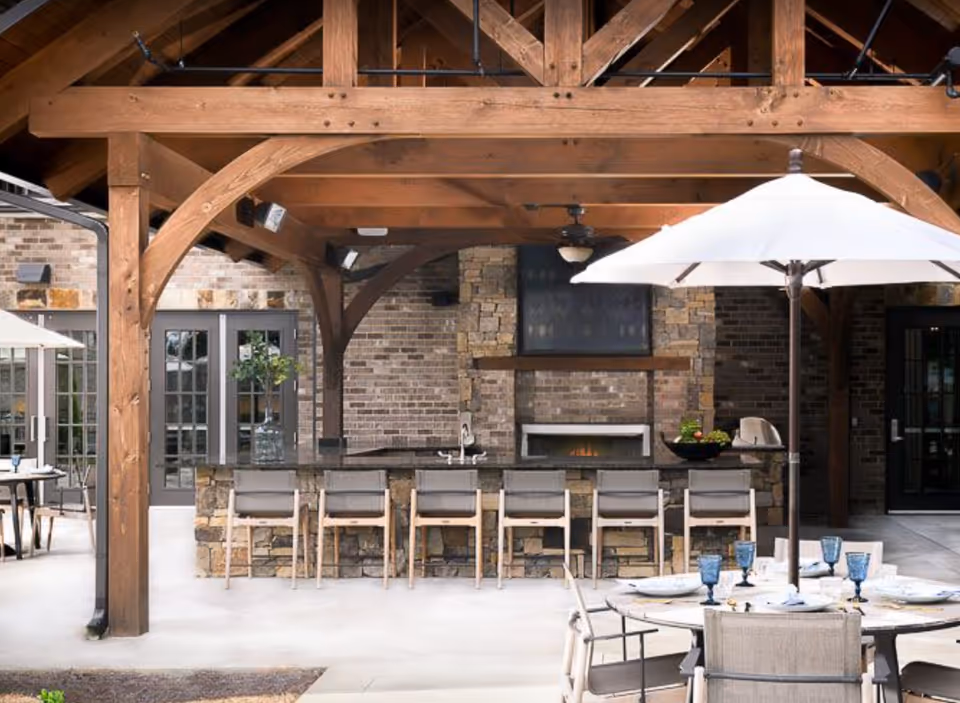 Outdoor covered patio area with a wooden beam structure, a stone bar with six bar stools, a fireplace built into a brick wall, and a round dining table with chairs and a large white umbrella.