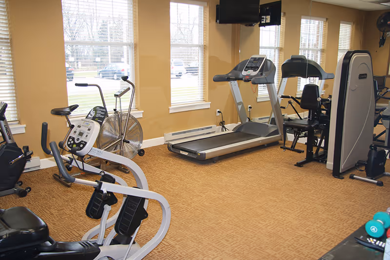 Small fitness room with treadmills, exercise bikes, a strength machine and free weights in front of large windows.