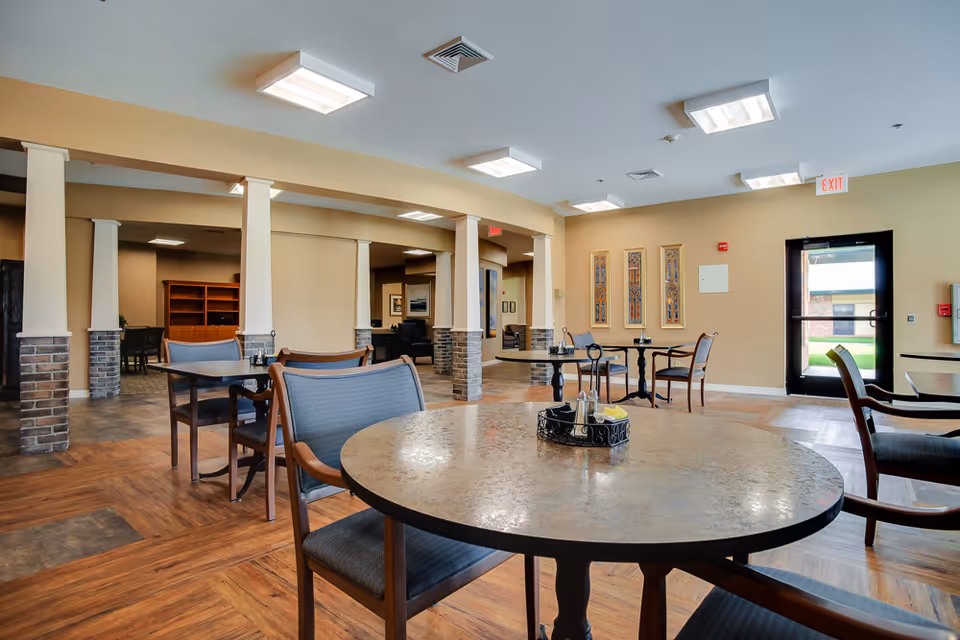 A spacious and well-lit dining area in a senior living facility with round tables and chairs arranged neatly. The room features beige walls, decorative wall art, and large windows near the exit door allowing natural light to enter. The floor has a wood and tile pattern, and the ceiling has multiple square light fixtures.