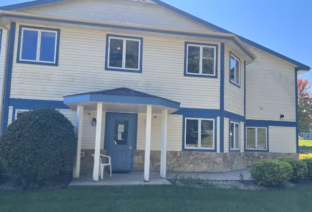 Exterior view of a two-story residential building with beige siding and blue trim. The entrance features a small covered porch with four white pillars and a blue door. There are several windows on both floors, and bushes are planted near the building. The sky is clear and blue.
