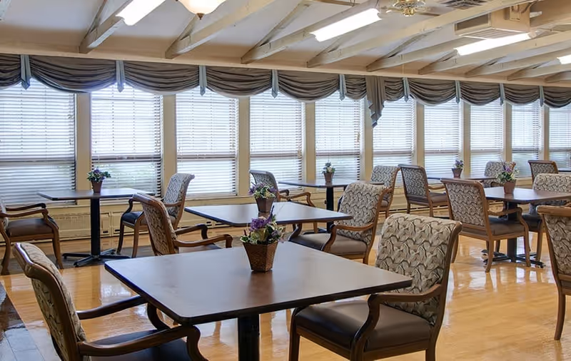 Bright dining room with multiple square tables and upholstered chairs, small potted flowers on each table, and a wall of windows with blinds and valances.