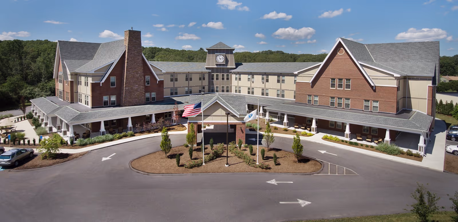 Front exterior view of The Residence at Valley Farm, a large senior living facility with a three-story building featuring brick and beige siding, a central clock tower, a covered entrance with two flagpoles displaying the American flag and another flag, surrounded by landscaped greenery and a circular driveway.