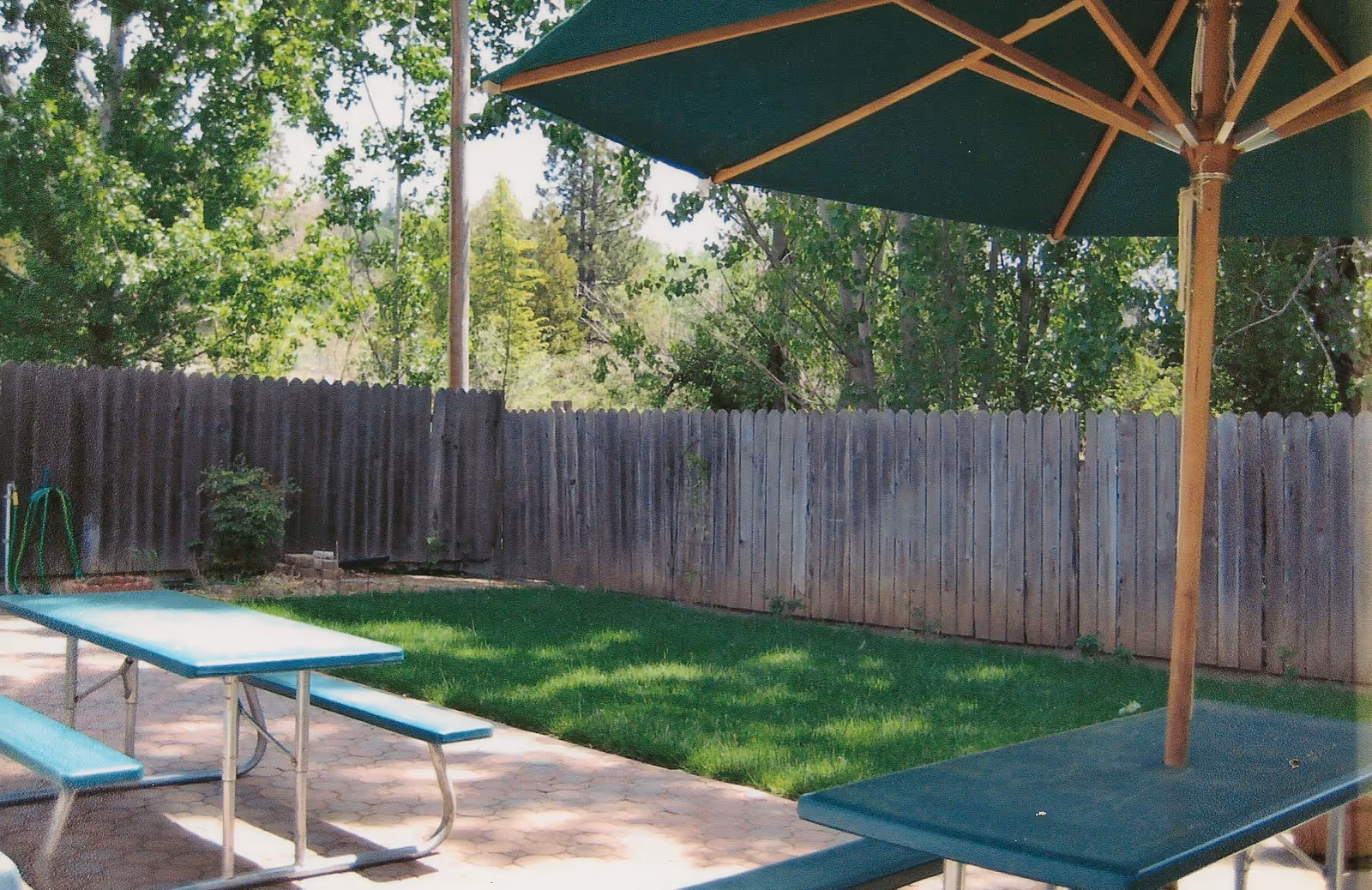 Outdoor patio area with green picnic tables and benches under a large green umbrella, surrounded by a wooden fence and trees in the background.