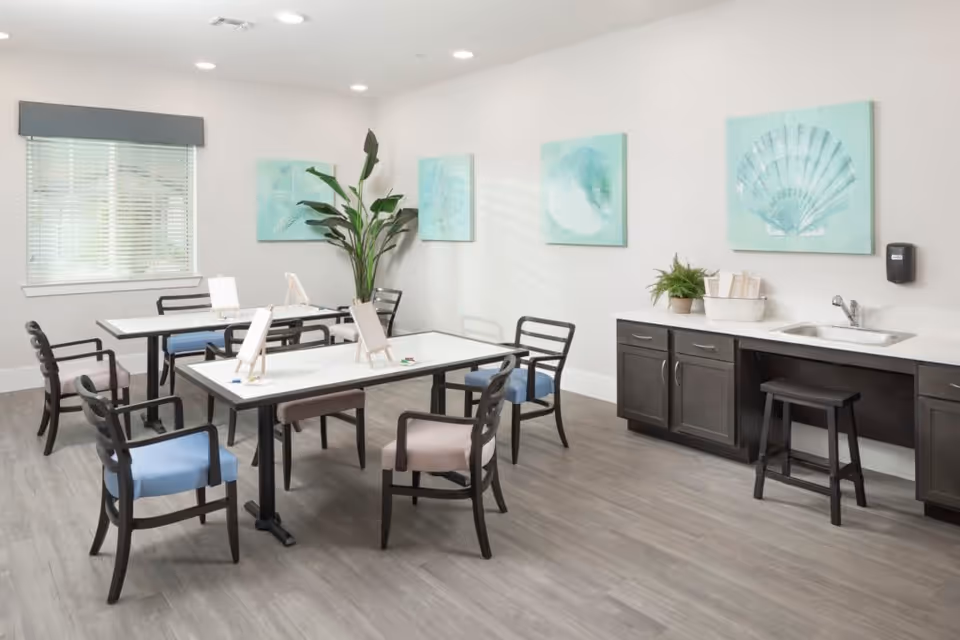 A bright and clean activity room with two rectangular tables surrounded by chairs with blue and beige cushions. Each table has small easels with paper and colored pencils for drawing or painting. The room features light-colored walls adorned with four sea-themed paintings, a large green plant in the corner, a window with blinds, and a countertop with a sink, cabinets, a stool, and a small plant.