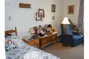 A cozy bedroom in a senior living facility with a bed covered in a floral bedspread and several stuffed animals on it. Next to the bed is a wooden chest with more stuffed animals and a small lamp. There is a blue upholstered armchair with a floor lamp beside it. The walls have a few framed pictures and a decorative mirror. A curtain covers a window on the right side.