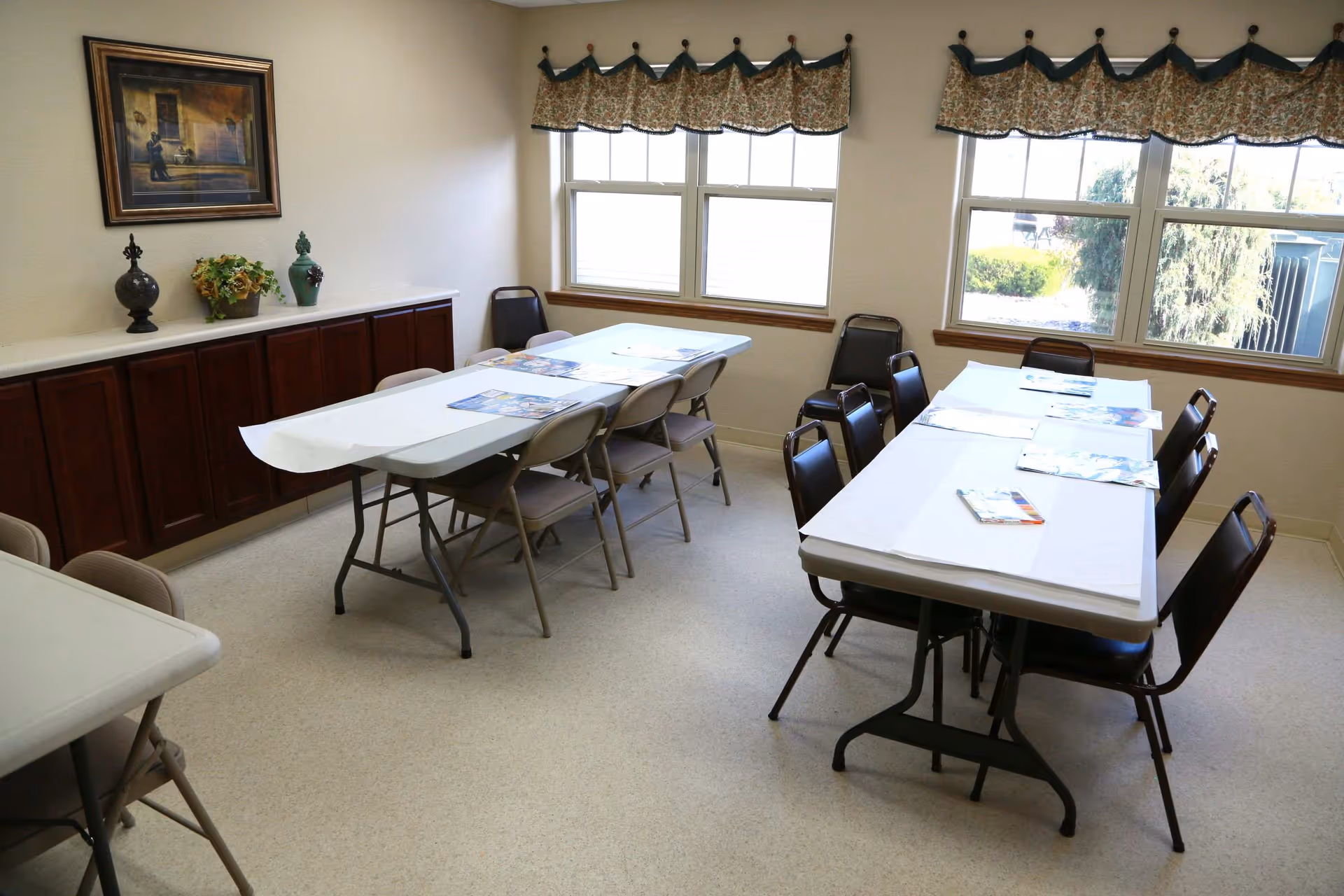 A bright room with two rectangular tables covered with white paper, surrounded by chairs. The room has three large windows with patterned valances, a long wooden cabinet against one wall with decorative items and a framed painting above it. The floor is light-colored and the walls are plain.