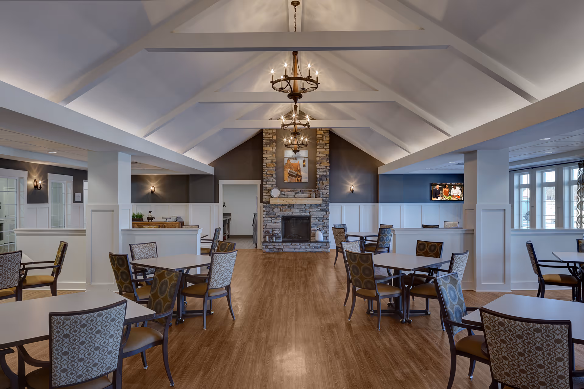 Spacious dining room with multiple tables and chairs arranged on a wooden floor. The room features a high vaulted ceiling with exposed beams and three chandeliers. At the far end, there is a stone fireplace with a framed picture above it. The walls are painted in a combination of dark and light colors with wainscoting, and there are large windows on the right side letting in natural light.