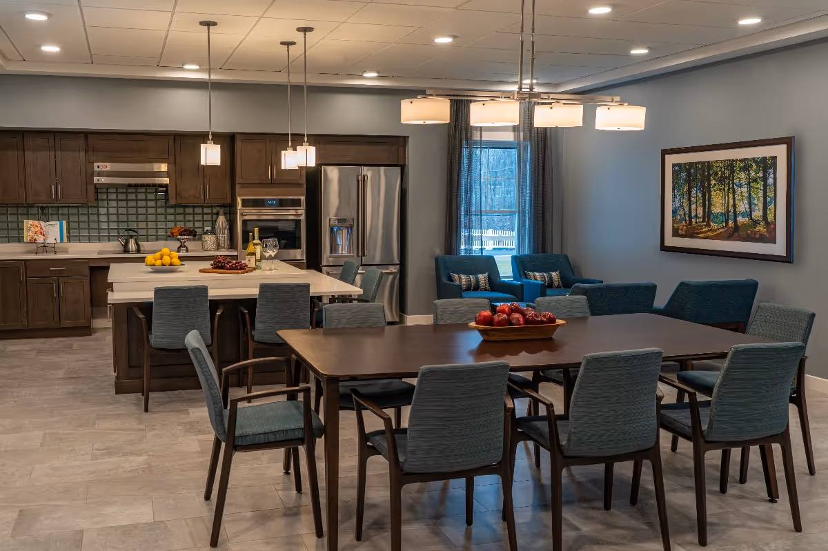 A modern open-concept kitchen and dining area featuring dark wood cabinets, stainless steel refrigerator, built-in oven, and a kitchen island with bar stools. The dining area has a large wooden table surrounded by eight blue upholstered chairs. In the background, there is a sitting area with blue armchairs and a window with curtains. The walls are painted light gray and decorated with a framed picture of a forest scene. Ceiling lights and pendant lights illuminate the space.