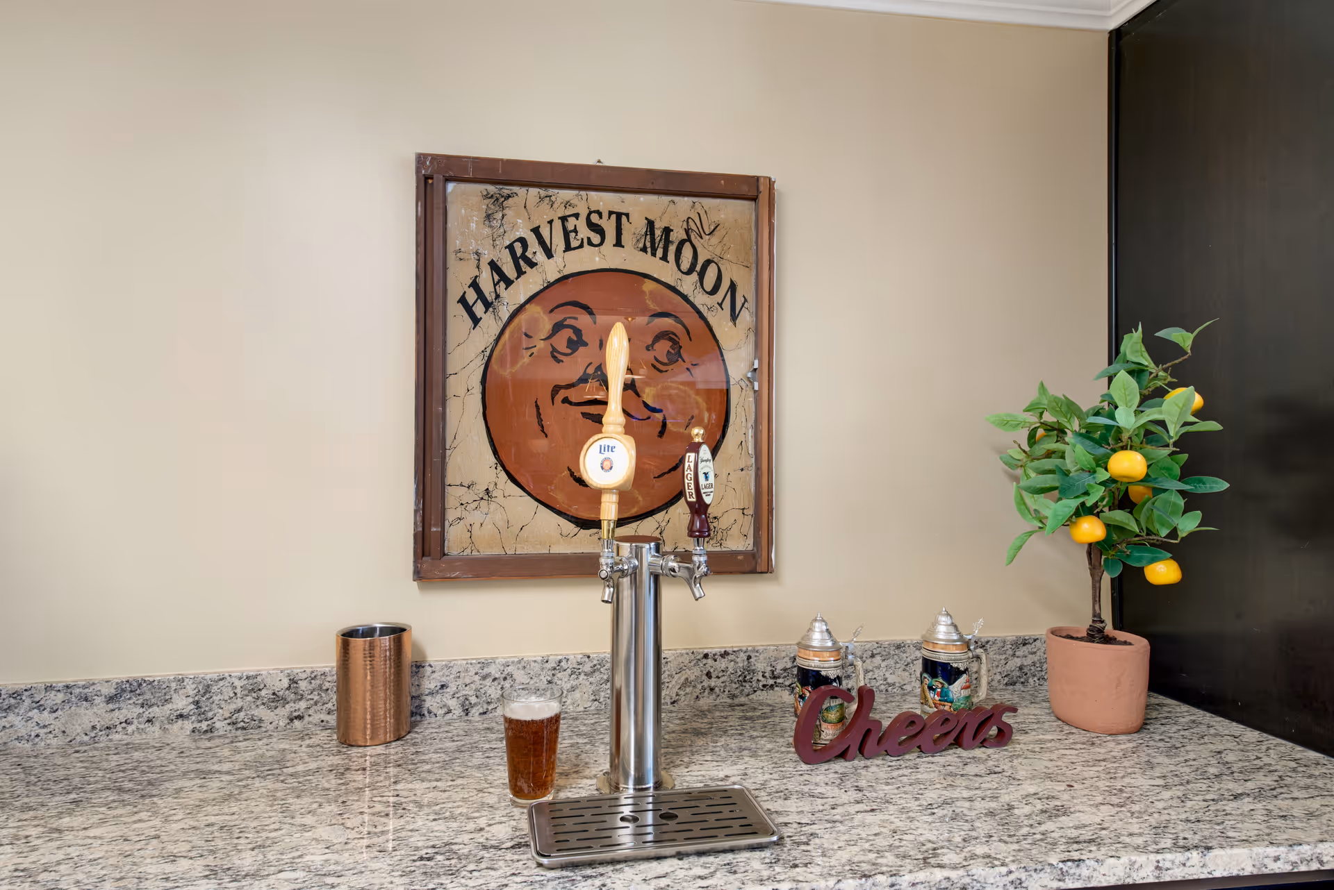 A countertop with a dual beer tap dispenser, a glass of beer, a copper cup, two decorative beer steins, a small potted plant with yellow fruit, and a wooden sign that says 'Cheers'. Above the counter is a framed vintage-style sign with a smiling orange moon face and the text 'HARVEST MOON'.
