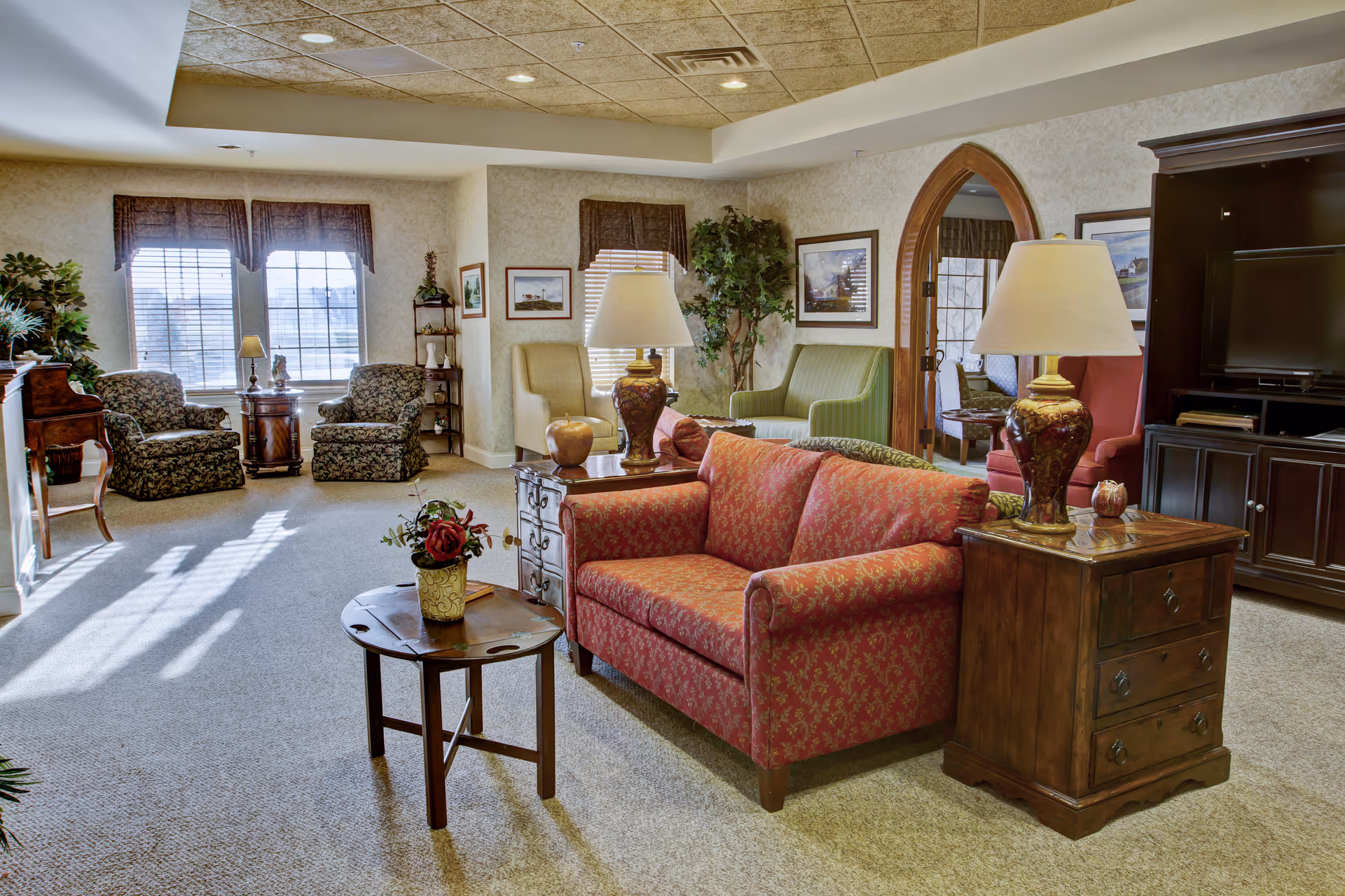 Sunlit cozy lounge in a senior living facility with patterned sofas and armchairs, side tables, lamps, and a TV cabinet.