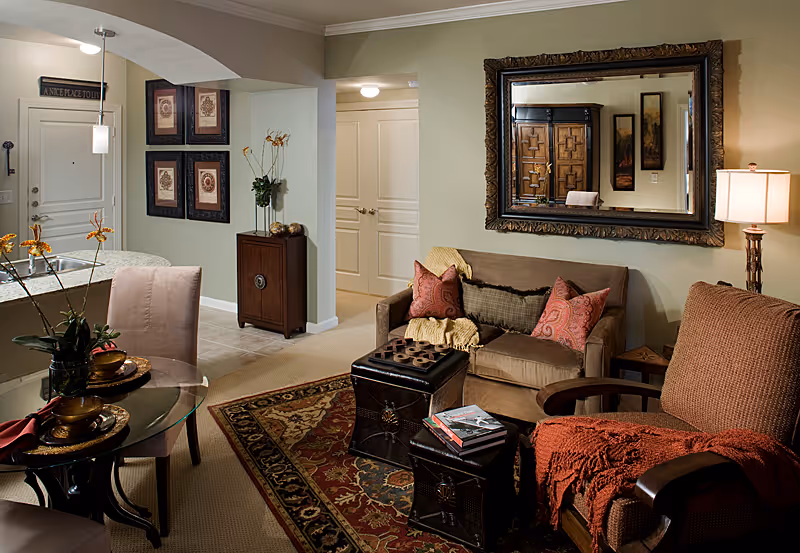 Cozy living room area with a brown sofa adorned with red and brown pillows and a yellow throw blanket. A large ornate mirror hangs on the wall above the sofa. In front of the sofa are two dark leather ottomans, one with books on top. To the right is a cushioned armchair with a red throw blanket. A floor lamp stands beside the armchair. To the left is a small round glass dining table with two upholstered chairs and decorative items on the table. The room has beige carpeting and a patterned area rug. The background shows a hallway with white double doors and framed artwork on the walls.