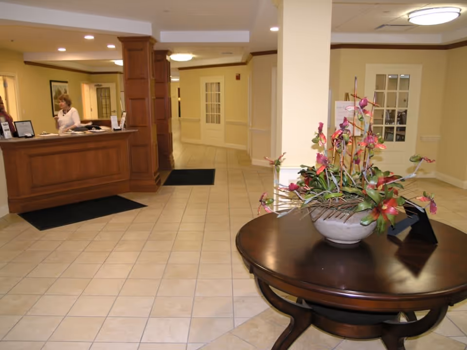 Reception area of a senior living facility with a wooden front desk on the left where a woman is standing, beige tiled floor, cream-colored walls, and a round wooden table in the foreground with a large floral arrangement. There are doors and hallways visible in the background.