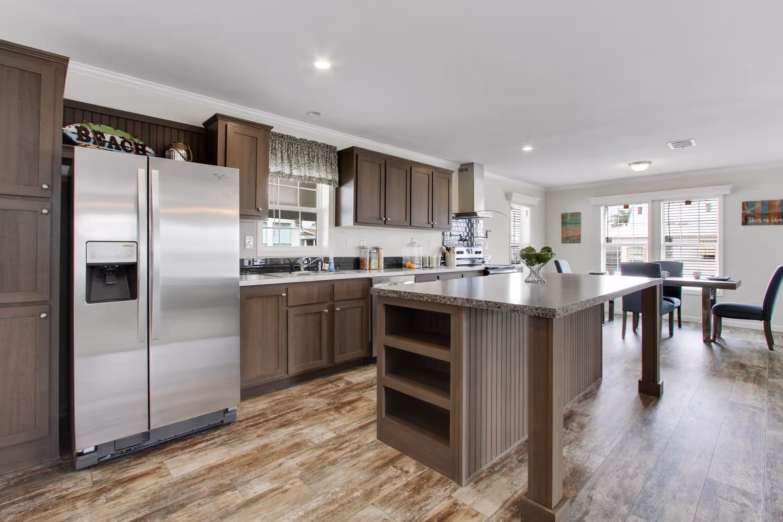 Modern kitchen with wooden cabinets, a large island with shelves, stainless steel refrigerator, stove with hood, and a dining area with table and chairs near windows with blinds.