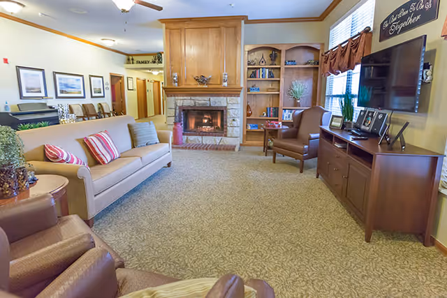A cozy living room in an assisted living facility featuring a beige sofa with striped pillows, two brown armchairs, a wooden fireplace with a stone base, built-in wooden bookshelves, a flat-screen TV on a wooden cabinet, and large windows with brown curtains letting in natural light.