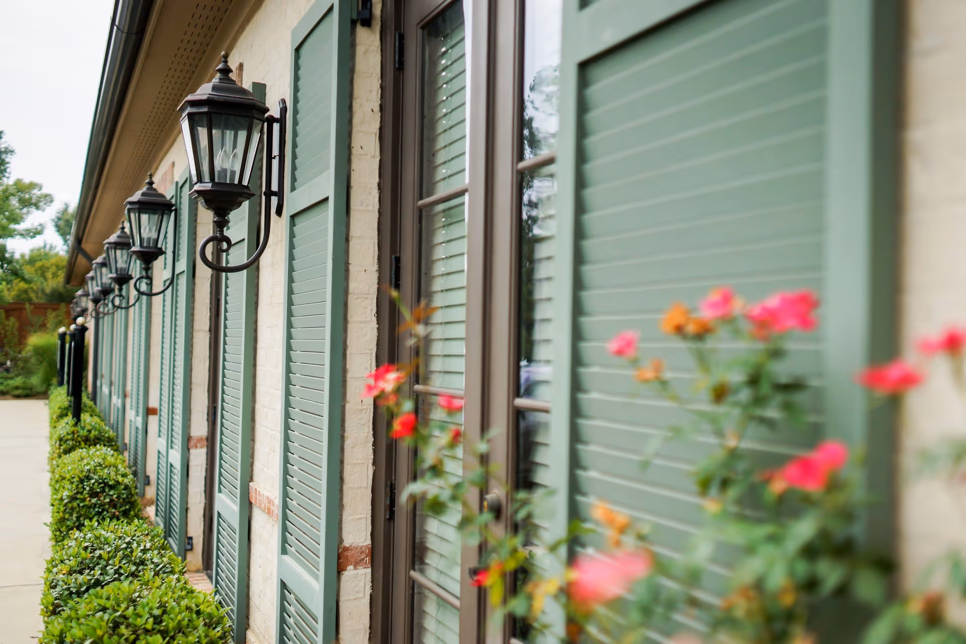 Exterior view of a building wall with green shutters on windows and black lantern-style wall lights. There are neatly trimmed bushes along the sidewalk and some pink and orange flowers in the foreground.