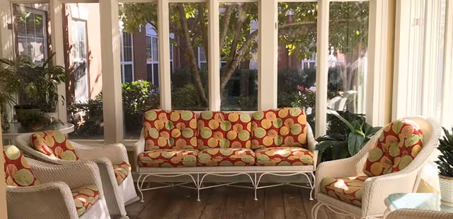 A sunlit indoor seating area with white wicker furniture including a sofa and three armchairs, all with cushions featuring a red and yellow apple pattern. Large windows surround the space, showing greenery and trees outside.