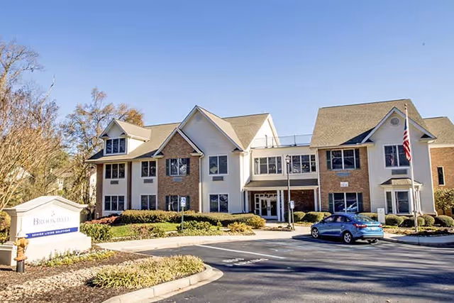 Exterior view of a two-story senior living facility building with a mix of brick and light-colored siding. The building has multiple windows, a main entrance with glass doors, and an American flag on a flagpole to the right. There is a parking lot in front with a blue car parked and landscaped greenery around the building. A sign near the entrance reads 'Brookdale Senior Living Solutions.'