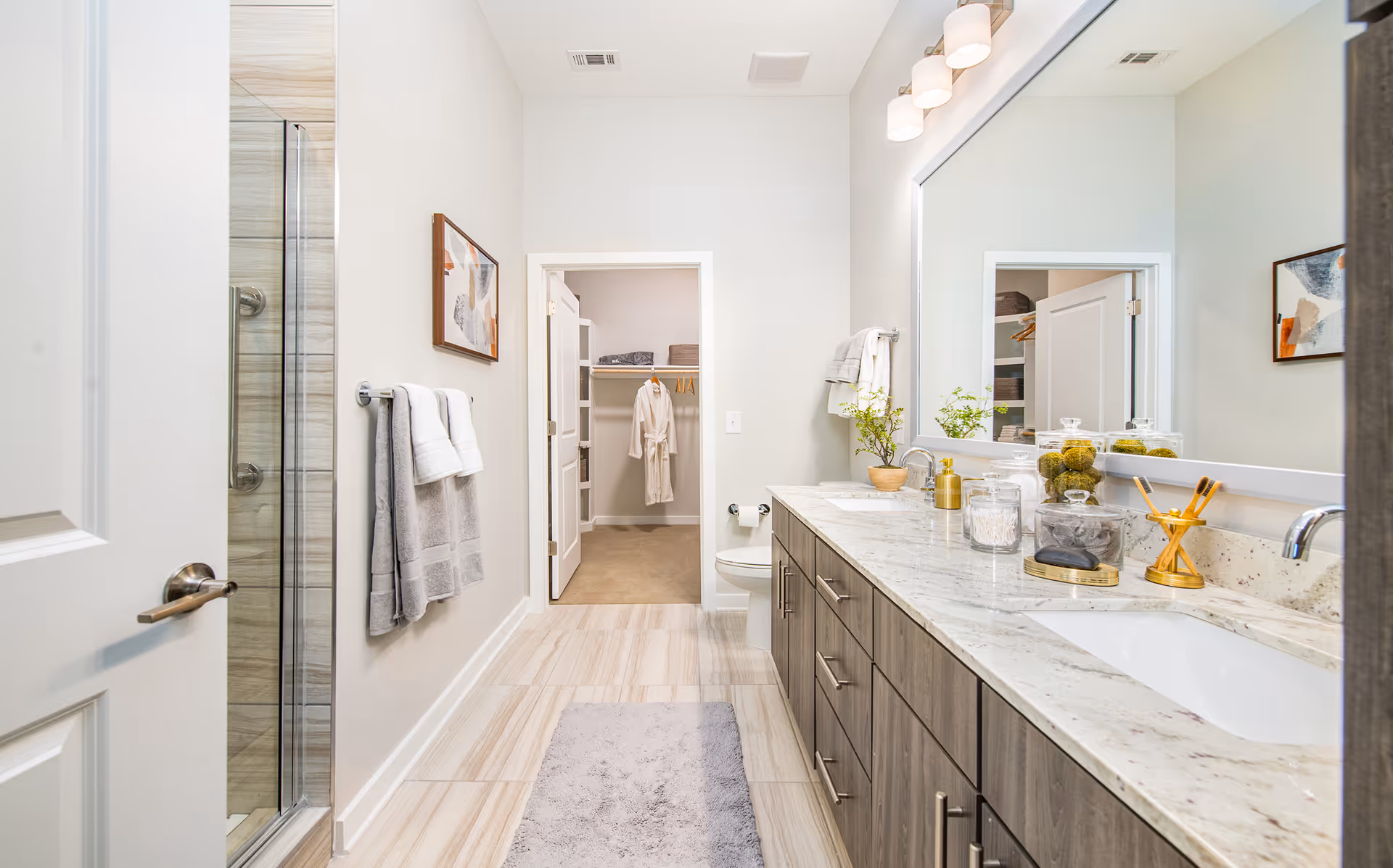 Bright modern bathroom with a double-sink marble vanity, large mirror, glass shower, towels on a rack, and a walk-in closet visible through a doorway.