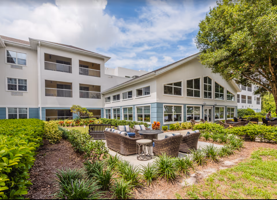 Outdoor seating area with wicker chairs and a table surrounded by greenery and plants in front of a multi-story building with balconies and large windows under a partly cloudy sky.