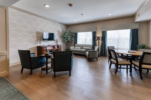 A well-lit living room area with wooden flooring featuring a sofa, armchairs, a flat-screen TV mounted on the wall above a wooden cabinet, a round dining table with chairs, large windows with curtains, and a floor lamp.