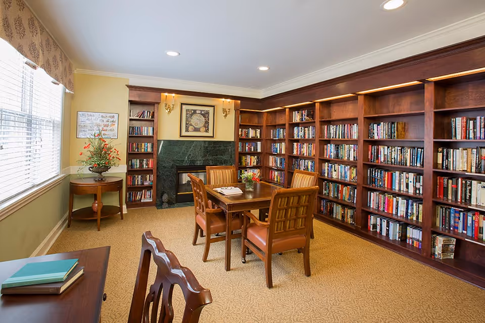 A cozy library/reading room with floor-to-ceiling bookshelves, a central table and chairs, a fireplace, and a large window.