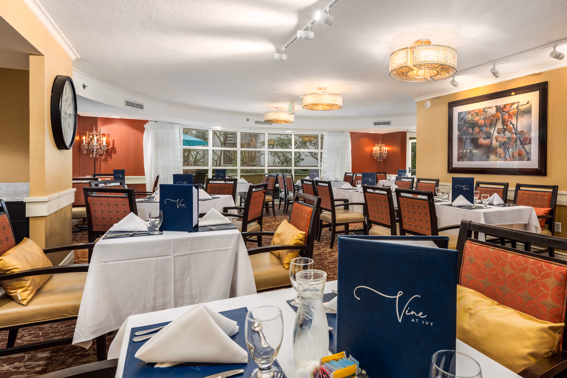 A well-lit dining room with multiple tables covered in white tablecloths, each set with folded white napkins, glassware, and blue menus labeled 'Vine at Ivy'. The room features cushioned chairs with red patterned upholstery and yellow accent pillows. Large windows with sheer white curtains allow natural light to enter, and there is a large framed artwork on one wall. The ceiling has modern light fixtures and the walls are painted in warm tones.
