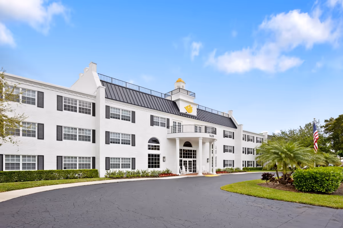 Exterior front view of a three-story white brick senior living facility building with black window shutters and a black metal roof. The entrance features a covered portico with white columns and a circular driveway. There are well-maintained shrubs, palm trees, and an American flag on the right side under a partly cloudy blue sky.