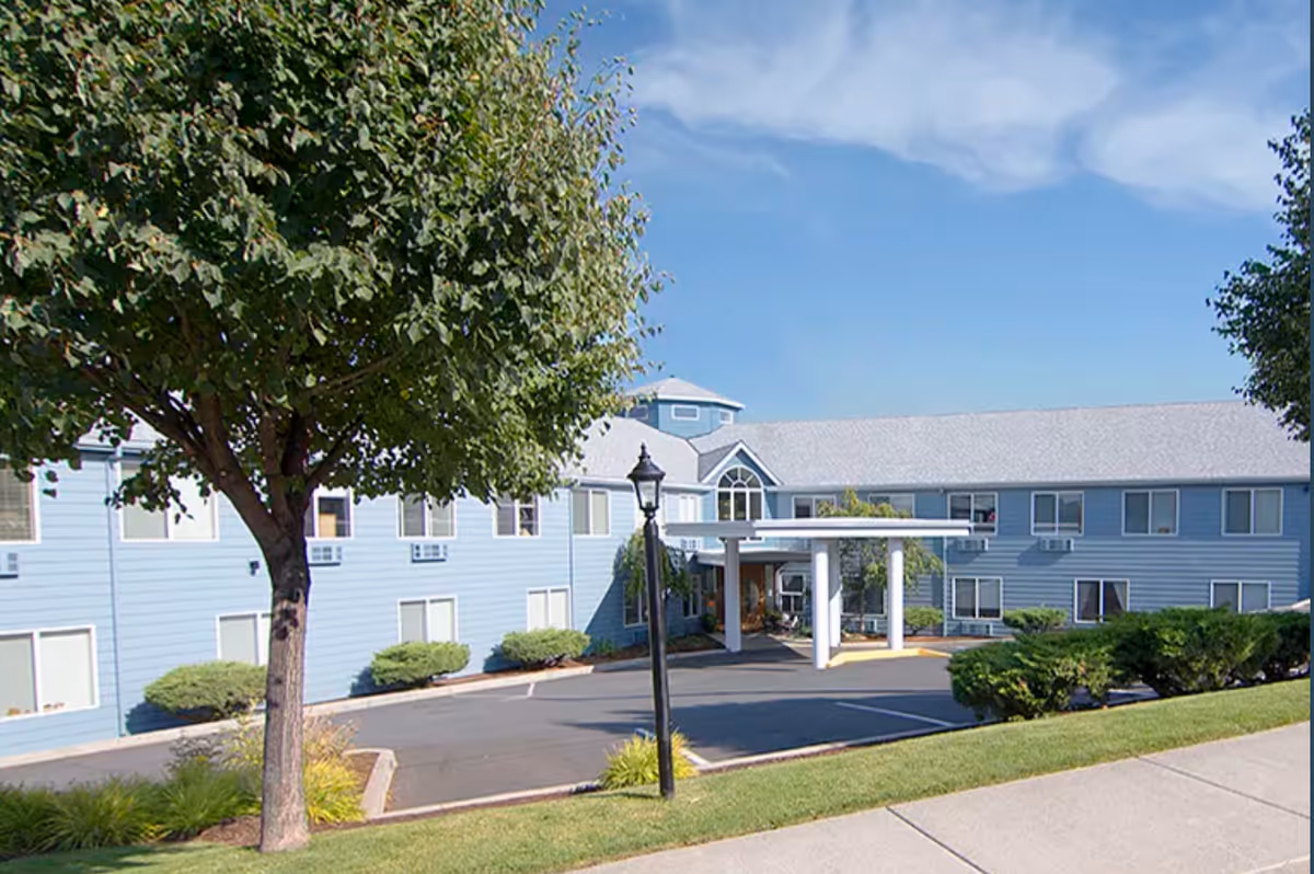 Exterior view of a two-story blue building with white trim, a covered entrance supported by white columns, surrounded by green bushes and trees under a blue sky with some clouds.
