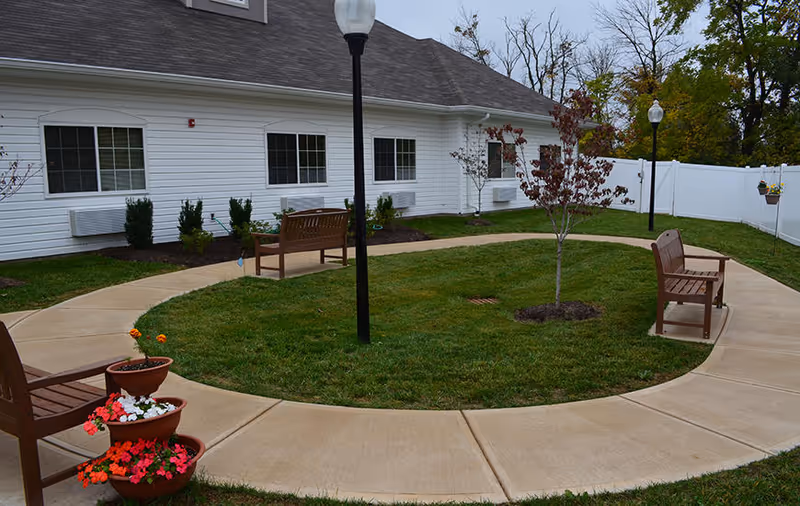 Outdoor courtyard area with a circular concrete walkway surrounding a grassy patch with small trees and flower pots. There are wooden benches placed along the walkway, and two black lamp posts providing lighting. The background shows a white building with multiple windows and a white fence enclosing the area.