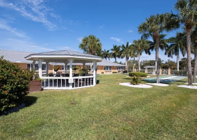 Outdoor lawn area with a white gazebo, palm trees, and a single-story building under a blue sky