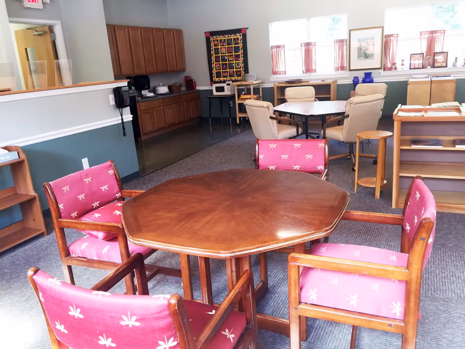 Interior room with a wooden octagonal table surrounded by four wooden chairs with pink cushions featuring white dragonfly patterns. In the background, there is a kitchenette area with wooden cabinets, a microwave on a small table, and a second table with four beige cushioned chairs. The room has windows with pink curtains, a framed picture on the wall, and various shelves and small furniture pieces.