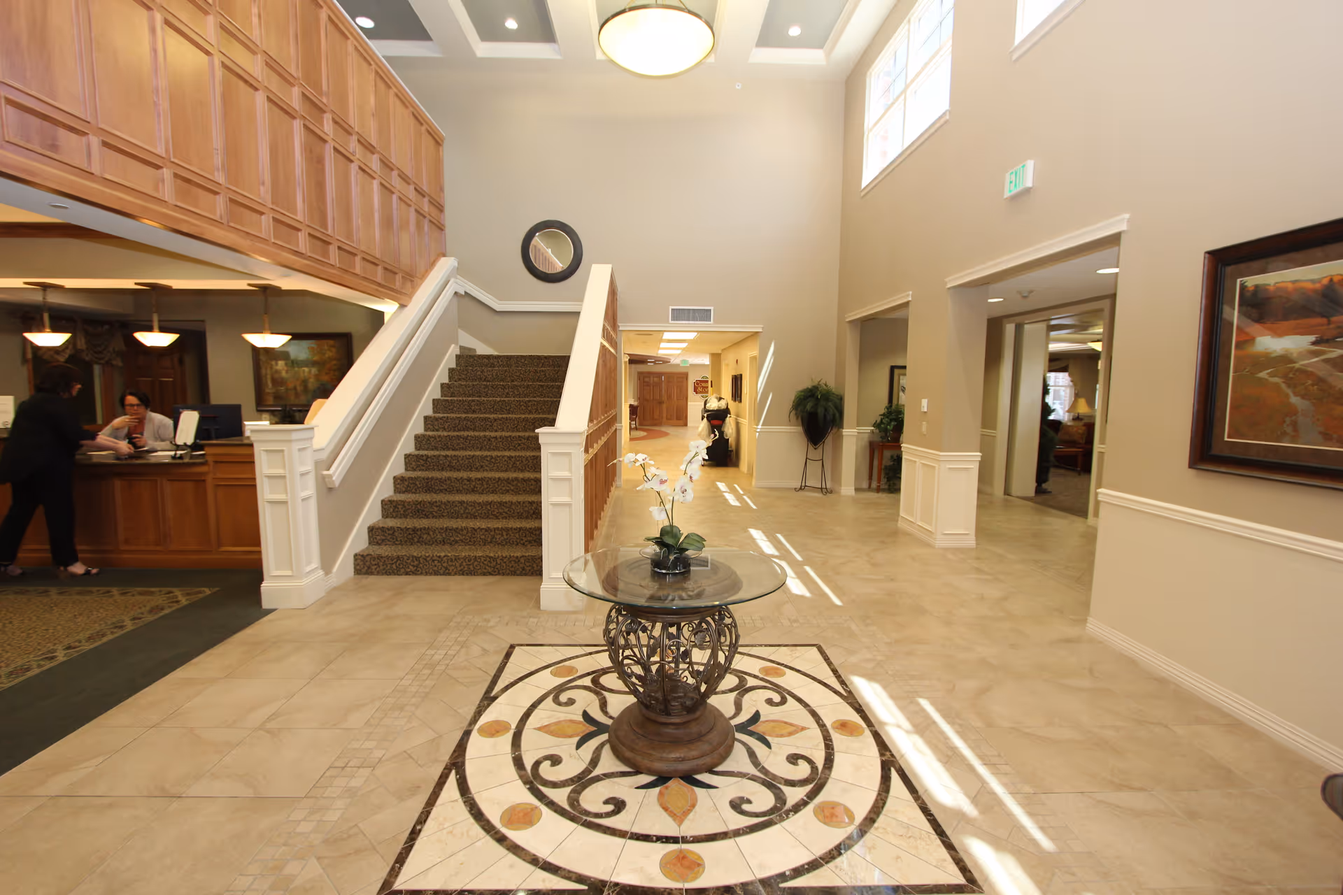 Interior view of a senior living facility lobby with a decorative round glass table holding a flower arrangement in the center. To the left, there is a reception desk with two women interacting. A carpeted staircase with wooden paneling leads to an upper floor. The walls are beige with white trim, and there are several windows near the ceiling letting in natural light. A hallway extends straight ahead with plants and artwork on the walls.
