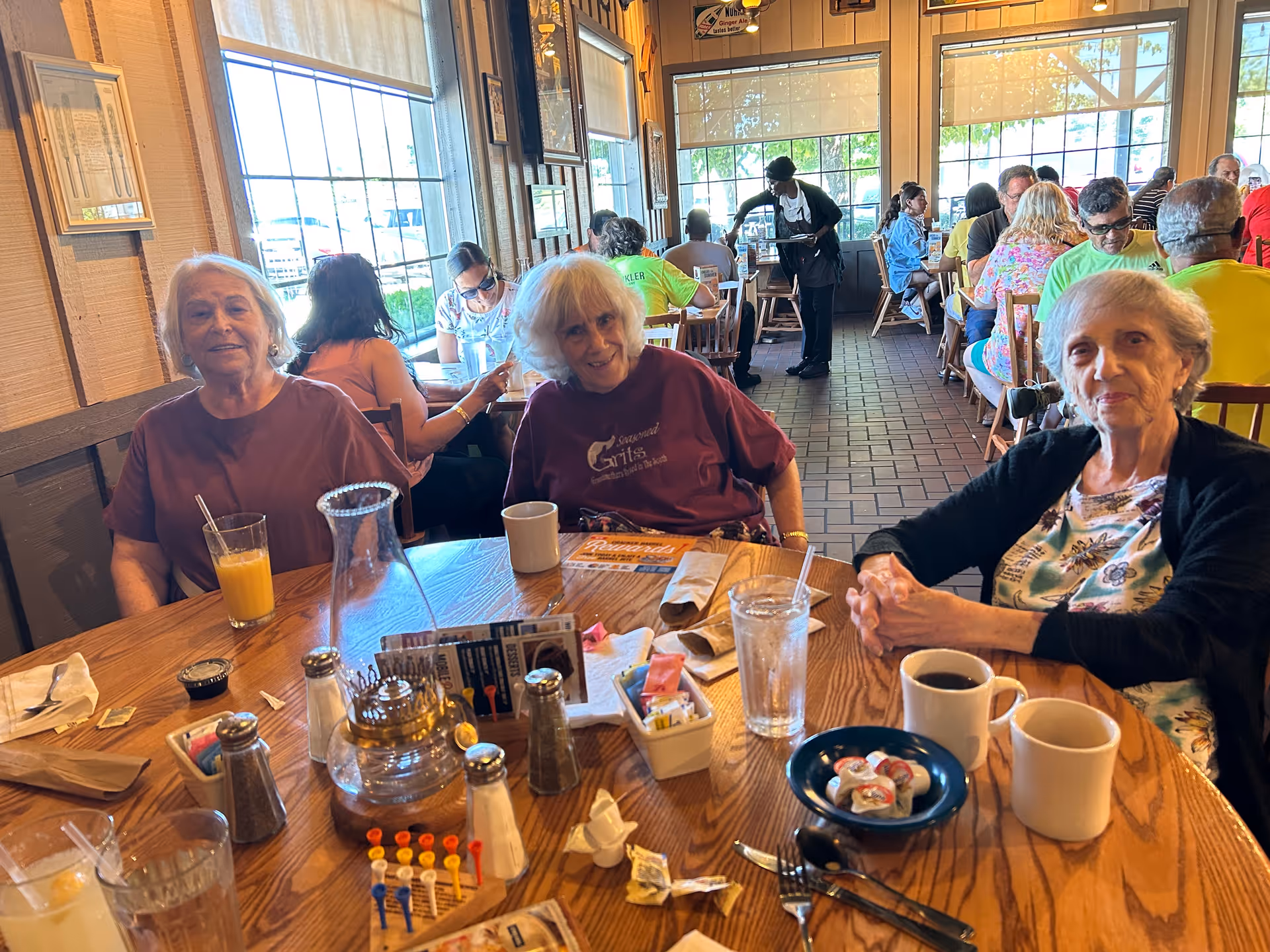 Three elderly women sitting at a wooden restaurant table with coffee, juice, condiments and tableware, other diners and a server visible in the background.