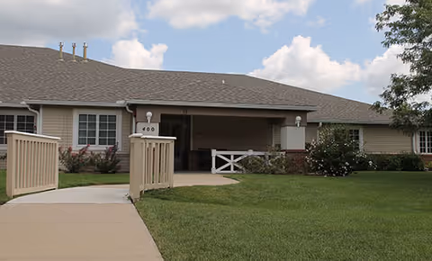 Exterior view of a single-story building with a gray roof and beige siding, featuring a covered entrance with white railings and a walkway leading up to it. The building is surrounded by green grass and some bushes, under a partly cloudy sky.