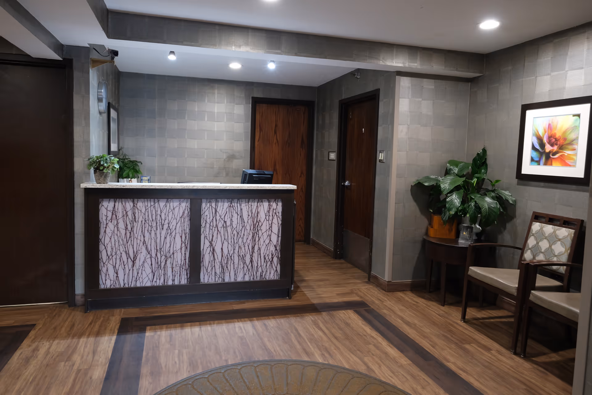Reception area with a front desk featuring a decorative panel, two wooden doors behind the desk, a small table with a green plant, two chairs with patterned cushions, and a colorful framed flower artwork on the wall.