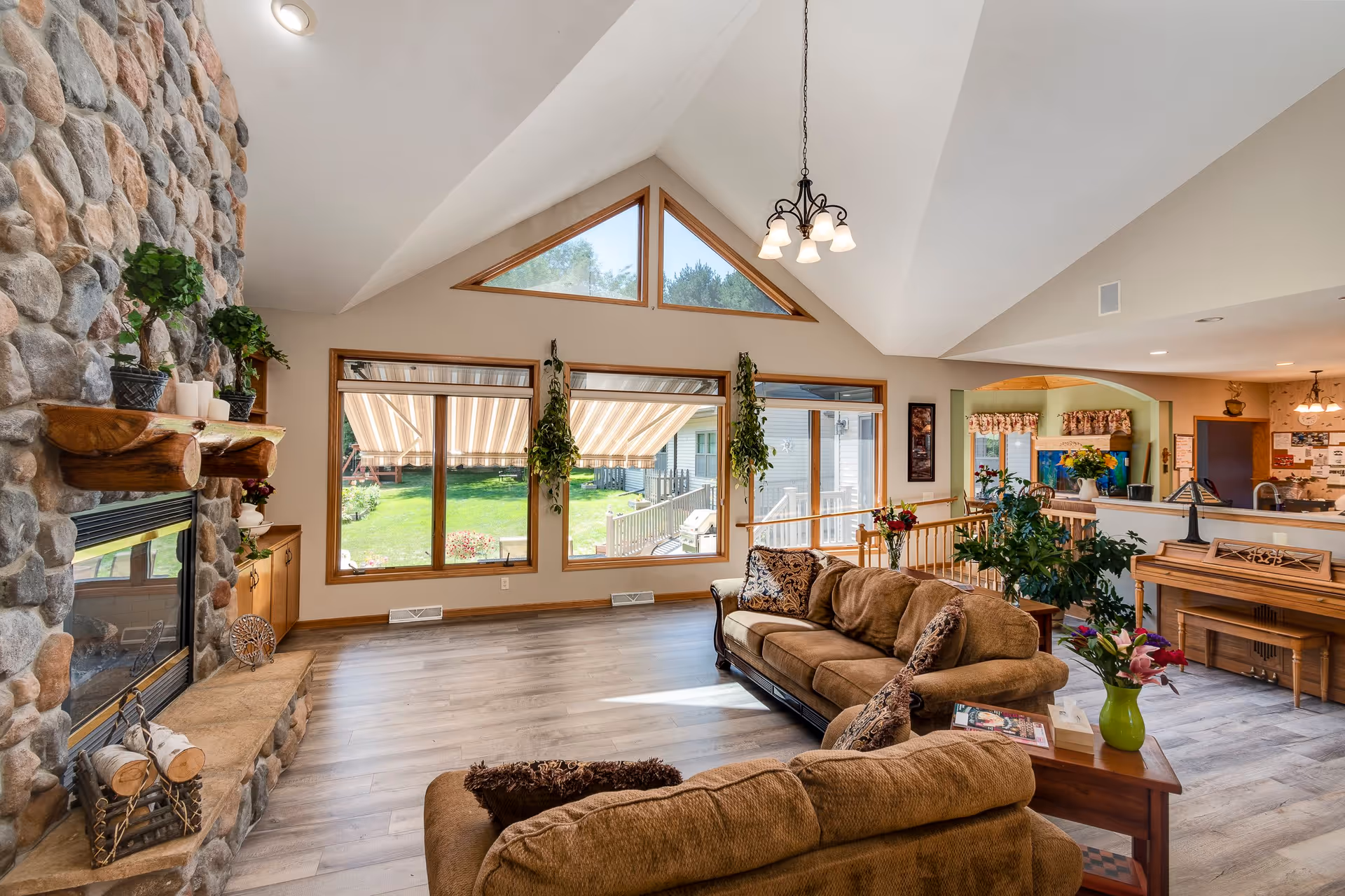 Spacious senior living facility common area with large windows letting in natural light, a stone fireplace with wooden mantel decorated with plants and candles, two brown sofas with patterned cushions, a wooden piano, and various plants and flowers. The room has high vaulted ceilings and light wood flooring.