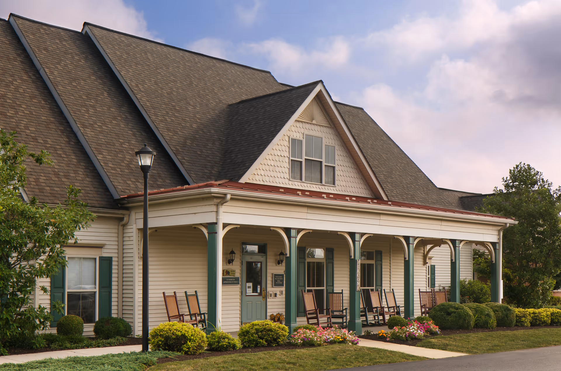 Front entrance of a cottage-style senior living building with a covered porch, several rocking chairs, and landscaped shrubs.