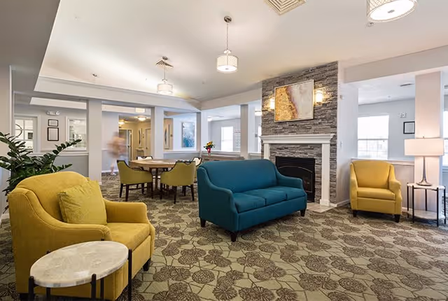 A bright and spacious living room area in an assisted living facility featuring a teal sofa, two yellow armchairs, a stone fireplace with a painting above it, a round table with chairs in the background, and patterned carpet flooring.