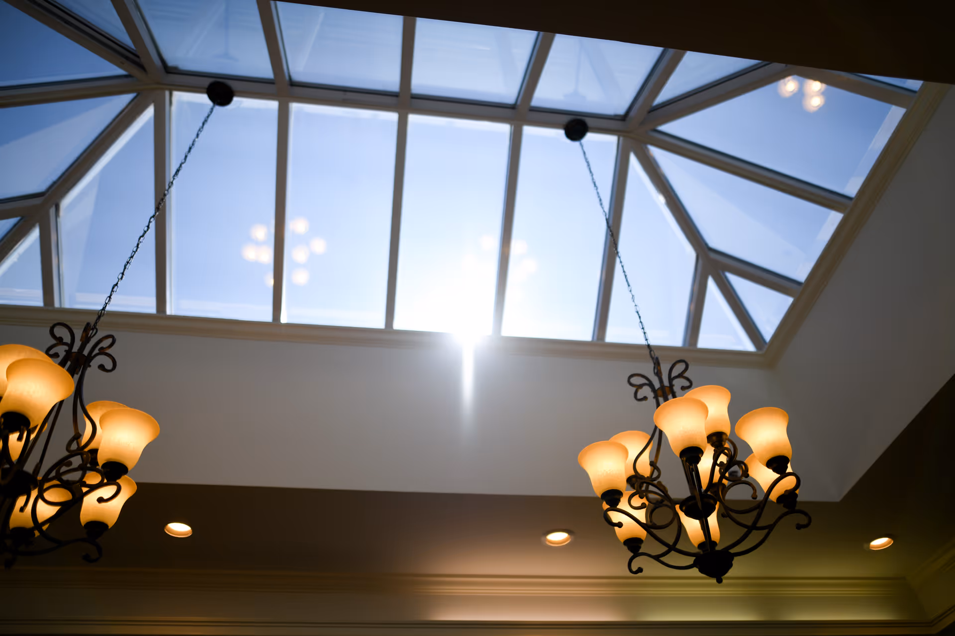 Skylight ceiling with two ornate hanging chandeliers and recessed lights in an interior space.